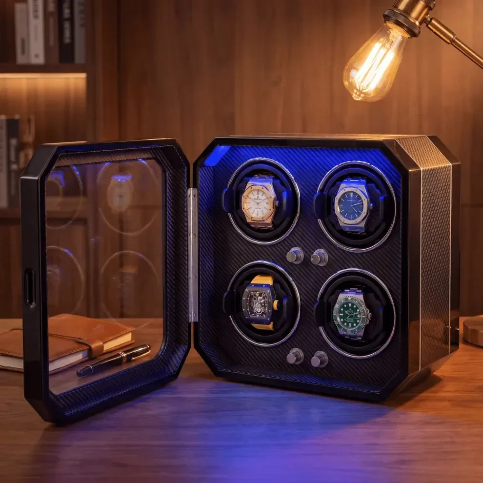 Black carbon fibre watch winder for 4 watches with blue LED lighting and glazed door, placed on a wooden desk.