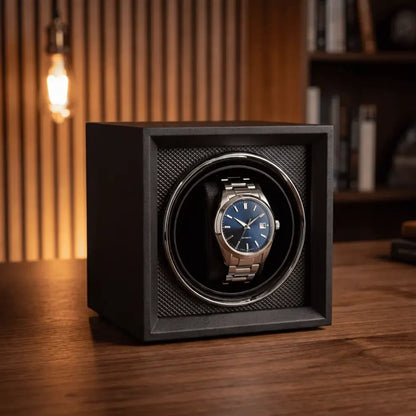 Black cube watch winder in wood and flannel with a blue-dial automatic watch on a wooden desk.