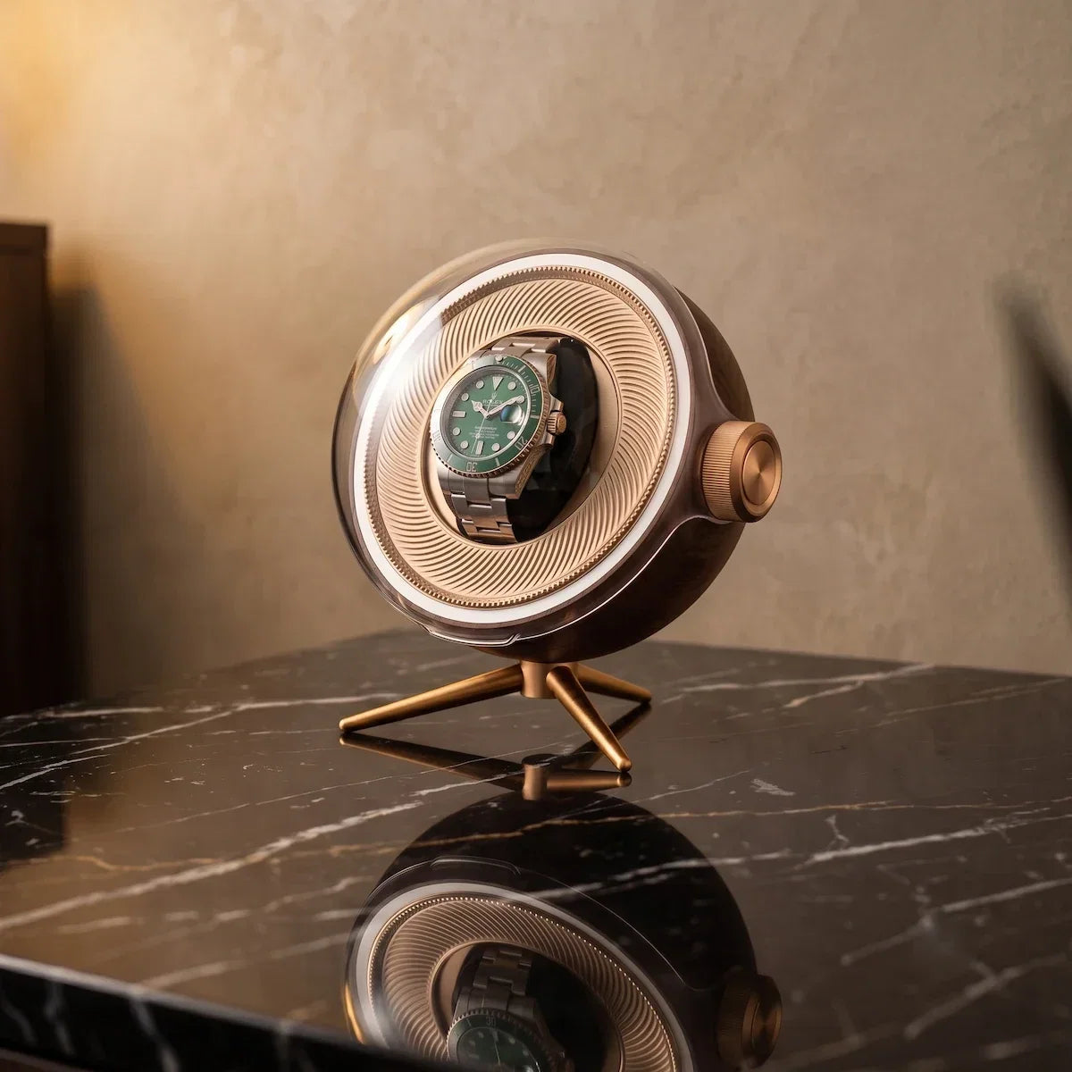 Tripode Solo watch winder with a spherical design in carbon colour and gold stand on a black marble table.